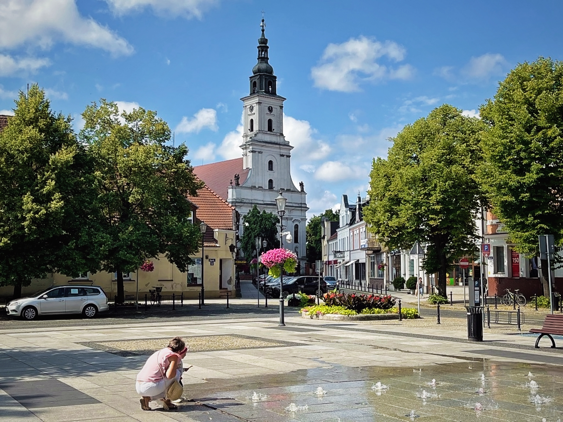 Plac miejski z fontanną w centrum, na którym widać osobę pochyloną nad strumieniami wody. W tle znajduje się wysoka wieża kościelna, otoczona drzewami i zadbanymi budynkami. Kwiaty w wiszących donicach oraz czyste, niebieskie niebo nadają scenerii pogodnej i spokojnej atmosfery, tworząc urokliwe, miejskie otoczenie.