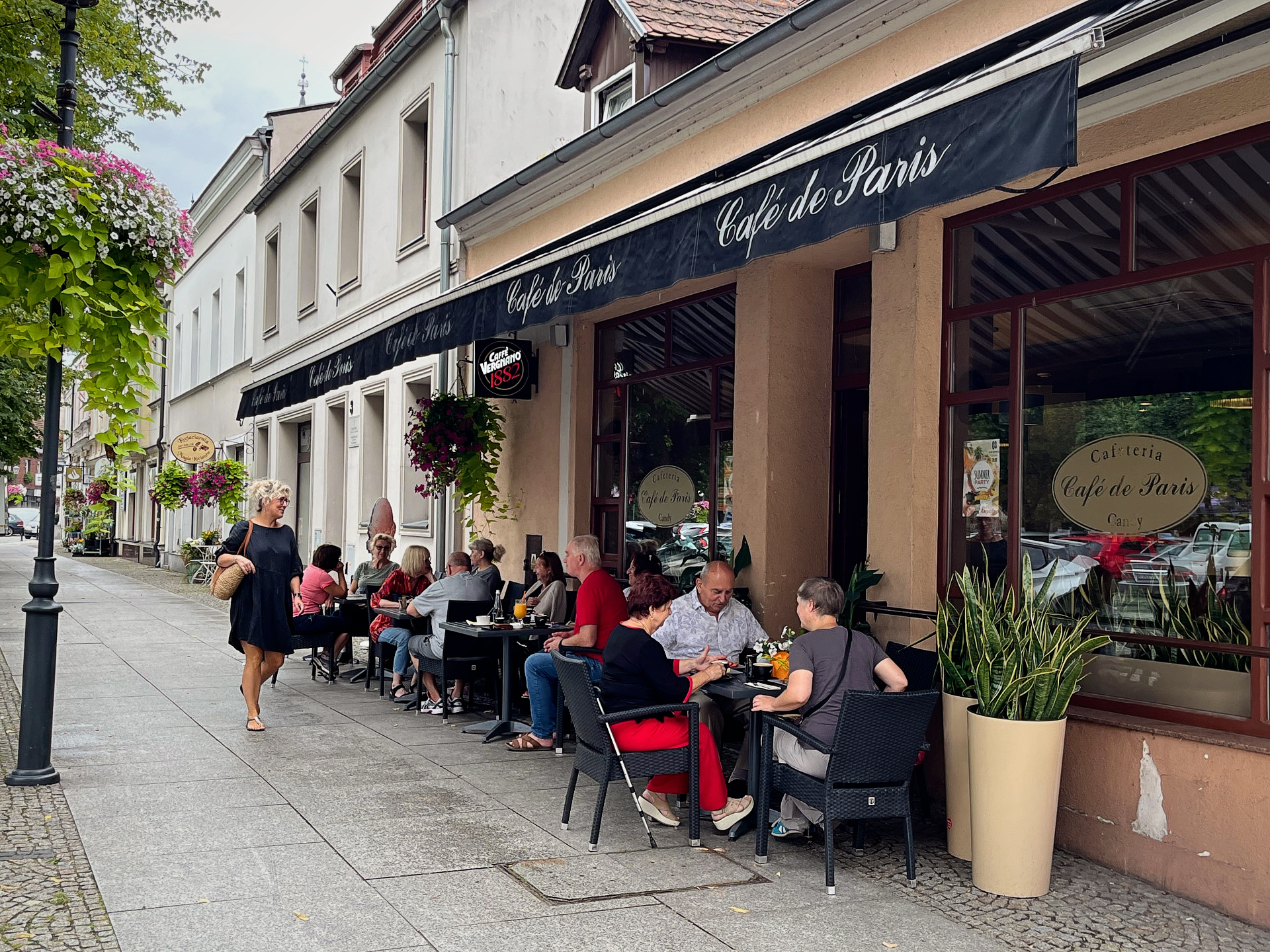 Ulica z kawiarnianym ogródkiem „Cafe de Paris”, gdzie przy stolikach na zewnątrz siedzą ludzie, rozmawiając i spożywając posiłki. Po lewej stronie widać chodnik ozdobiony kwiatami w wiszących donicach, a w tle kamienice. Atmosfera jest spokojna i przyjazna, a ludzie cieszą się czasem spędzonym na świeżym powietrzu w miejskim otoczeniu.