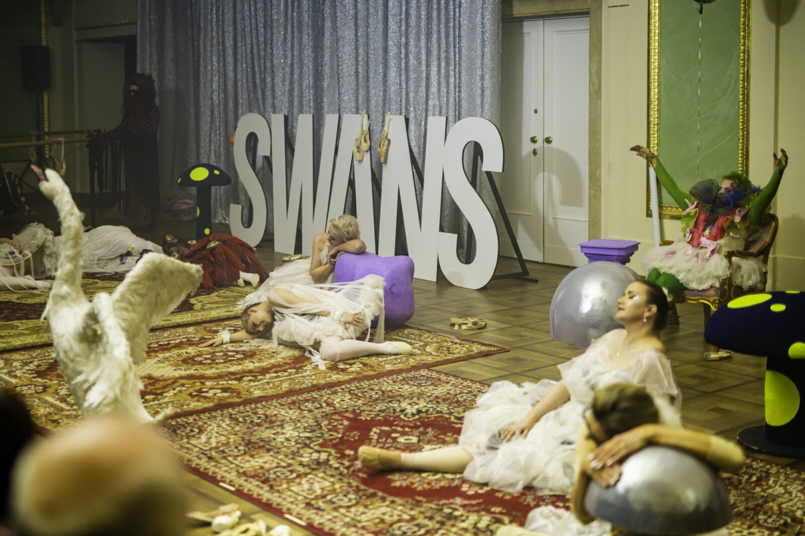 Tancerki w białych, zwiewnych kostiumach w zastygłych pozach wykonują scenę ze spektaklu „Łabędzie” w Teatrze Wielkim w Poznaniu. Część z nich leży na podłodze, inne są w półleżących pozach, jakby pogrążone we śnie. Scenę wypełniają czerwone dywany, duże litery tworzące napis „SWANS” oraz fantazyjne elementy scenografii przypominające grzyby i łabędzia.